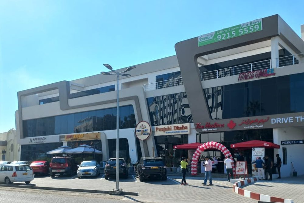 The Corner from the road in Al Khuwair: shopfronts at street level under two upper floors, with a drive-through lane.